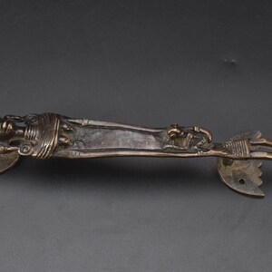 May include: A dark bronze-toned metal door handle featuring a stylized, elongated human figure stretched along the length of the bar. The handle includes two decorative mounting plates at each end, each with a single pre-drilled screw hole.