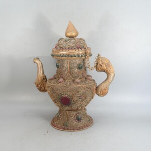 May include: A metallic teapot featuring intricate silver-toned filigree wirework, a dragon-shaped handle, and a spout. The vessel is adorned with embedded dark red and green cabochon stones, a pointed lid, and a small chain connecting the lid to the body.