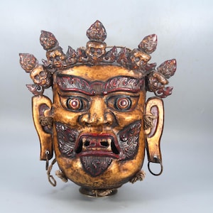 Puede incluir: Una máscara antigua y detallada con una expresión feroz. La máscara es principalmente dorada con detalles rojos alrededor de los ojos, la boca y elementos decorativos. Presenta una estructura similar a una corona y detalles en las orejas.