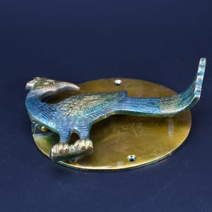 May include: A brass door knocker featuring a bird figure with a blue and green patina finish mounted on a circular brass plate. The bird is depicted in profile with textured wings and a long tail, attached to a polished round base with two visible screw holes.