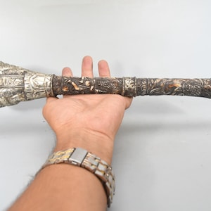 Puede incluir: Una maza ceremonial de metal larga y cilíndrica con una cabeza acampanada ornamentada y un eje oscuro y desgastado cubierto de intrincados relieves de figuras y patrones, acabado con una sección superior metálica de tono plateado.