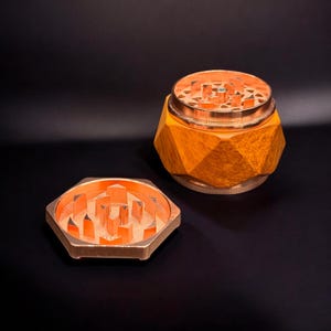 Pode incluir: Um moedor facetado de cor cobre com uma tampa correspondente. O moedor est&aacute; aberto, revelando o interior com dentes angulares e afiados. A base &eacute; de cor laranja quente e o fundo &eacute; preto.