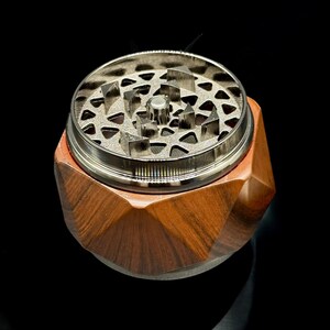 Pode incluir: Um moedor de ervas de madeira com um design geom&eacute;trico. O moedor tem um topo de metal prateado com dentes afiados para moer. A base &eacute; feita de madeira marrom escura com um design facetado. O moedor est&aacute; em uma superf&iacute;cie preta.
