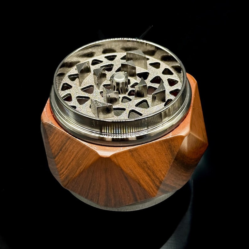 Pode incluir: Um moedor de ervas de madeira com um design geom&eacute;trico. O moedor tem um topo de metal prateado com dentes afiados para moer. A base &eacute; feita de madeira marrom escura com um design facetado. O moedor est&aacute; em uma superf&iacute;cie preta.