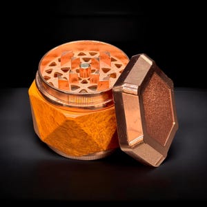Pode incluir: Um moedor de ervas de cor cobre com um design geom&eacute;trico. O moedor est&aacute; aberto, revelando o mecanismo de moagem. A tampa tem formato de diamante e repousa ao lado da base. A base tem uma apar&ecirc;ncia texturizada e facetada.