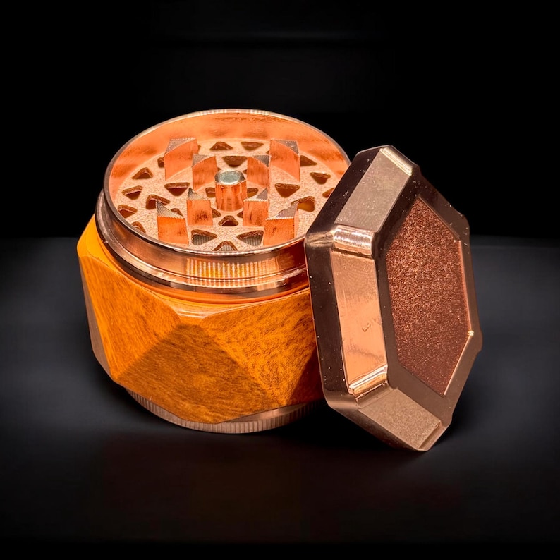 Pode incluir: Um moedor de ervas de cor cobre com um design geom&eacute;trico. O moedor est&aacute; aberto, revelando o mecanismo de moagem. A tampa tem formato de diamante e repousa ao lado da base. A base tem uma apar&ecirc;ncia texturizada e facetada.
