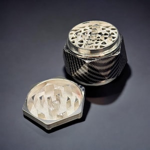 Pode incluir: Um moedor de ervas prateado e preto com um padr&atilde;o de fibra de carbono no corpo. O moedor &eacute; hexagonal com uma tampa e uma pe&ccedil;a inferior separada, ambas com dentes de moagem prateados. A tampa est&aacute; aberta.