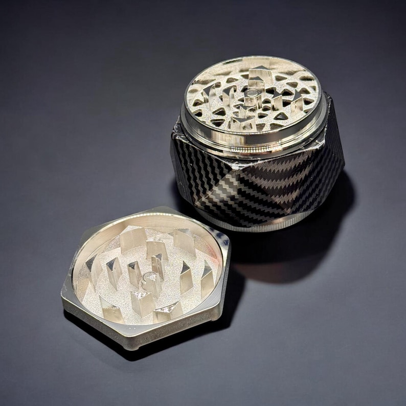 Pode incluir: Um moedor de ervas prateado e preto com um padr&atilde;o de fibra de carbono no corpo. O moedor &eacute; hexagonal com uma tampa e uma pe&ccedil;a inferior separada, ambas com dentes de moagem prateados. A tampa est&aacute; aberta.