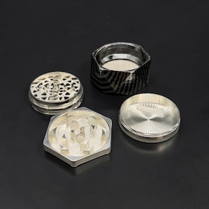 Pode incluir: Um moedor de ervas de metal prateado desmontado com uma parte superior com padr&atilde;o de fibra de carbono. O moedor est&aacute; em v&aacute;rias pe&ccedil;as, incluindo uma base hexagonal, uma parte superior com design e uma tampa.