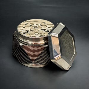 Pode incluir: Um moedor de ervas prateado e preto com um padr&atilde;o de fibra de carbono na base. A parte superior est&aacute; aberta, revelando os dentes de moagem. A tampa tem uma forma geom&eacute;trica. O moedor est&aacute; sobre uma superf&iacute;cie preta.
