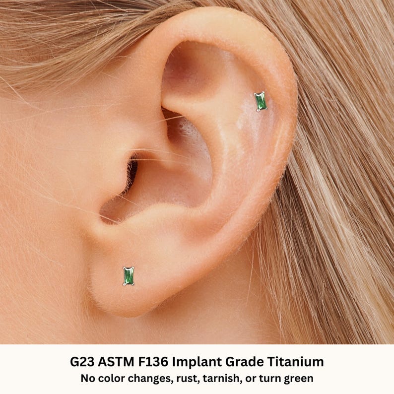 May include: Close-up of a person's ear with two small green gemstone earrings. The earrings are made of G23 ASTM F136 Implant Grade Titanium. The text "No color changes, rust, tarnish, or turn green" is below the image.