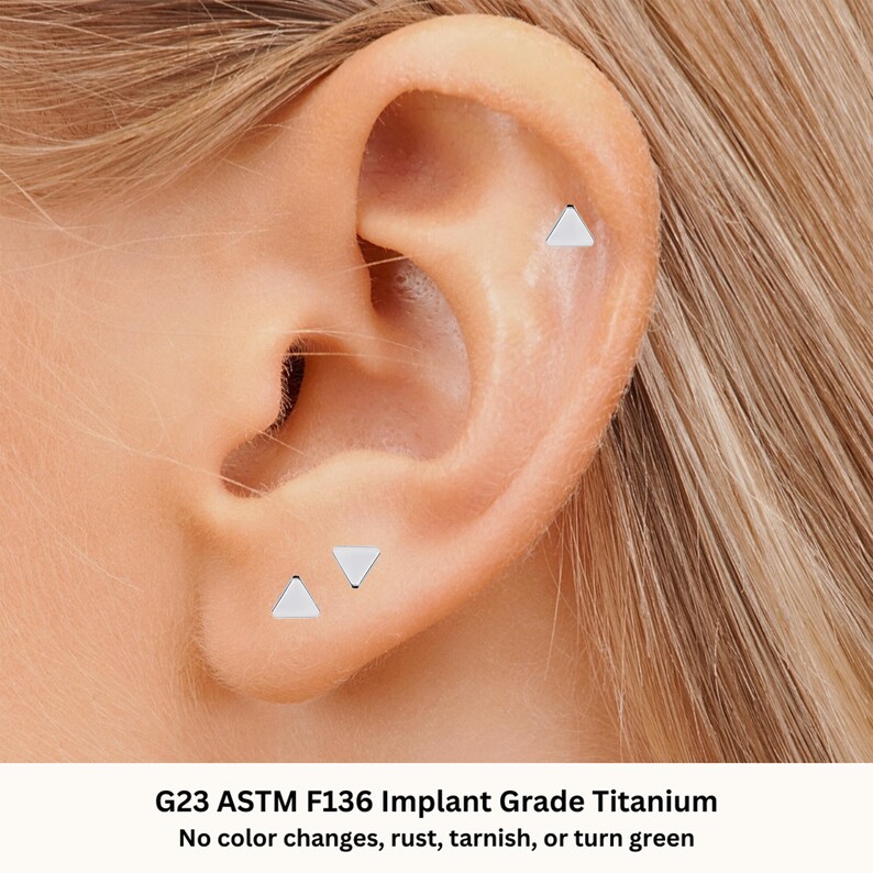 May include: Three small silver triangle earrings in a person's ear. The earrings are made of G23 ASTM F136 Implant Grade Titanium. The text "No color changes, rust, tarnish, or turn green" is below the image.