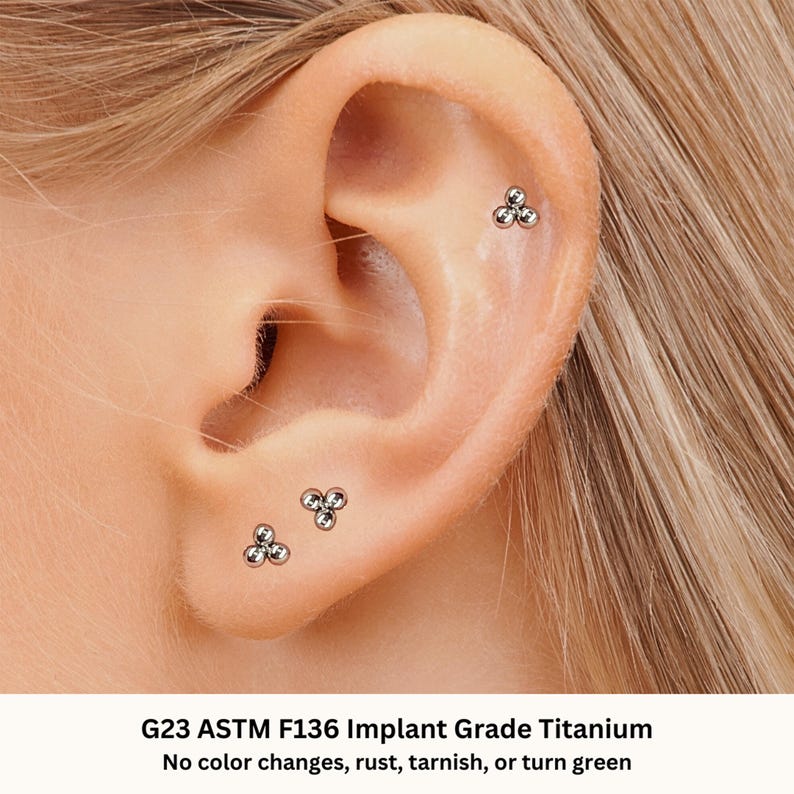 May include: Close-up of a person's ear with three small, silver, triple ball stud earrings. The earrings are made of G23 ASTM F136 Implant Grade Titanium. The text "G23 ASTM F136 Implant Grade Titanium" and "No color changes, rust, tarnish, or turn green" is visible below the image.
