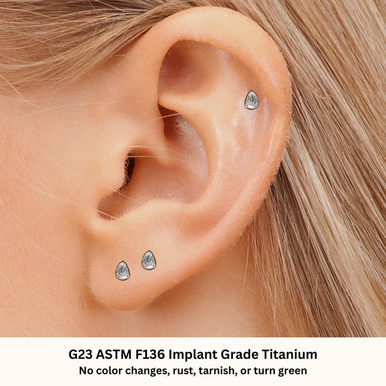 May include: Three small, clear, teardrop-shaped earrings in a person's ear. The earrings are made of G23 ASTM F136 Implant Grade Titanium. The text "G23 ASTM F136 Implant Grade Titanium" and "No color changes, rust, tarnish, or turn green" is below the image.