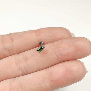 May include: A silver body piercing with a small, emerald green, rectangular gem.