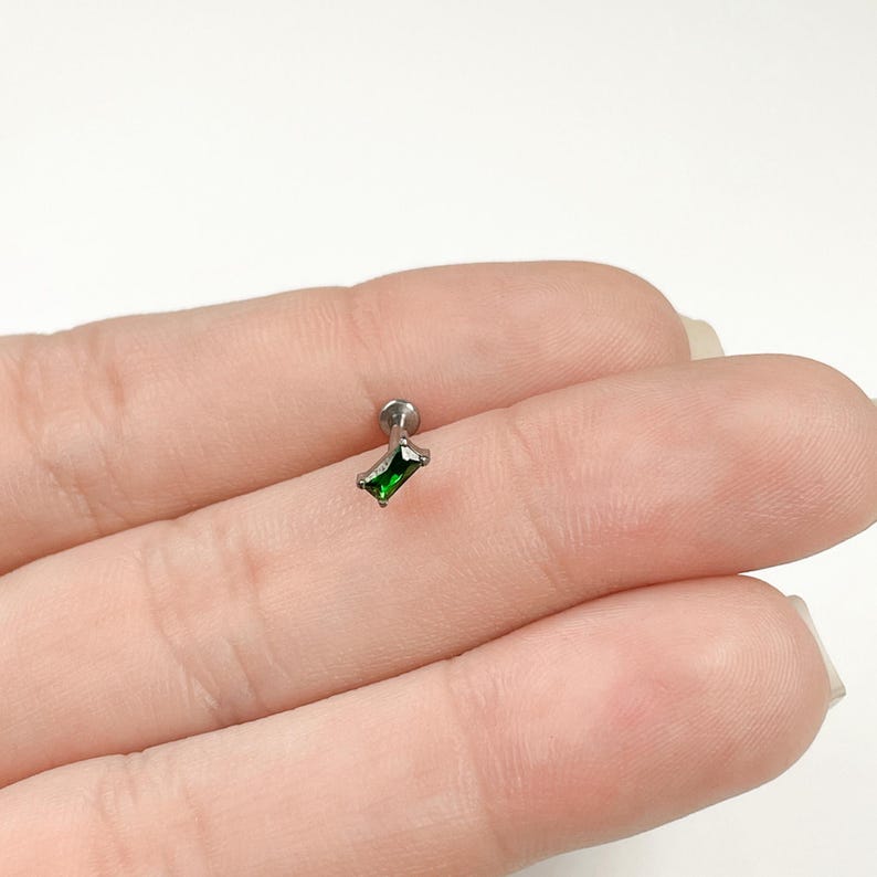 May include: A silver body piercing with a small, emerald green, rectangular gem.
