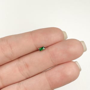 May include: A small, emerald green, rectangular-shaped gemstone set in a silver stud earring.