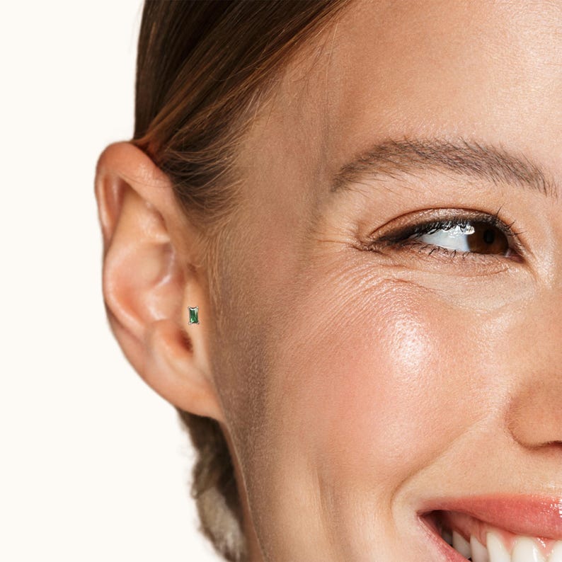 May include: A close-up of a person's ear with a small, emerald green gemstone stud earring. The earring is set in a gold setting.