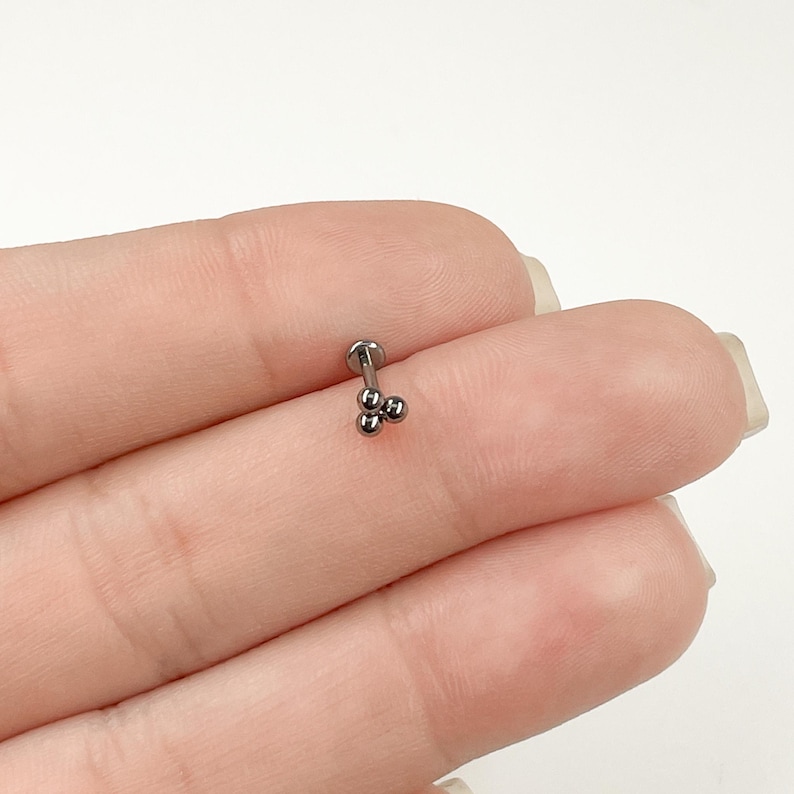 May include: A silver barbell with three small silver balls on the end. The barbell is a body piercing jewelry item.