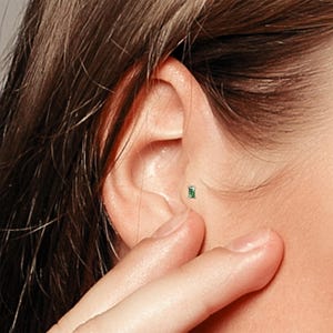 May include: A close-up of a person's ear with a small green gemstone stud earring. The earring is set in a silver or white gold setting.