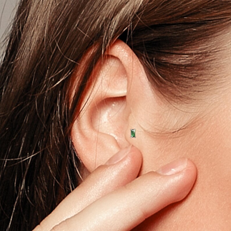 May include: A close-up of a person's ear with a small green gemstone stud earring. The earring is set in a silver or white gold setting.