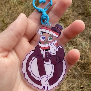 May include: A clear acrylic keychain with a cartoon character in a maid outfit and a top hat. The character has a wide grin, mismatched eyes, and white gloves. The keychain has a blue heart-shaped clasp.