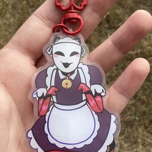 May include: A clear acrylic charm featuring a cartoon maid character with a white mask, purple dress, and red accents. The charm is attached to a red metal clasp.