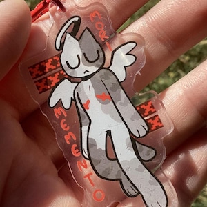 Puede incluir: Un llavero acrílico transparente con un gato de dibujos animados con alas de ángel. El gato es gris y blanco con detalles en rojo. El llavero tiene una cuerda roja y texto en rojo.