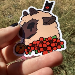 May include: A sticker featuring a cartoon lamb with a black crown and red eye. The lamb has a brown face and is surrounded by red flowers and a bell. The sticker is held up by a hand against a blurred green background.