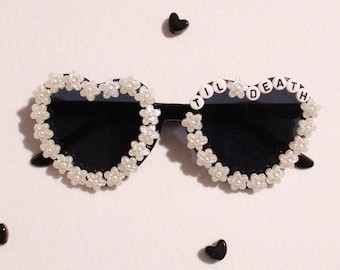 May include: Heart-shaped sunglasses with black lenses and a white floral frame. The phrase "TIL DEATH" is spelled out in white letter beads across the top. Small black heart-shaped beads are scattered around the sunglasses on a white background.