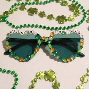 Peut inclure: Des lunettes de soleil vertes avec le texte "Kiss Me I'm Irish" et d&eacute;cor&eacute;es de strass verts, oranges et transparents. Les lunettes sont entour&eacute;es de colliers de perles vertes et de perles en forme de tr&egrave;fle.