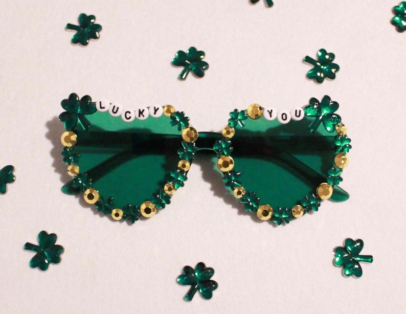 K&ouml;nnte beinhalten: Gr&uuml;ne Sonnenbrille mit Kleeblatt- und Goldperlenbesatz. Der Text "LUCKY YOU" ist &uuml;ber den Gl&auml;sern angebracht. Kleeblatt-Konfetti ist um die Brille herum verstreut.