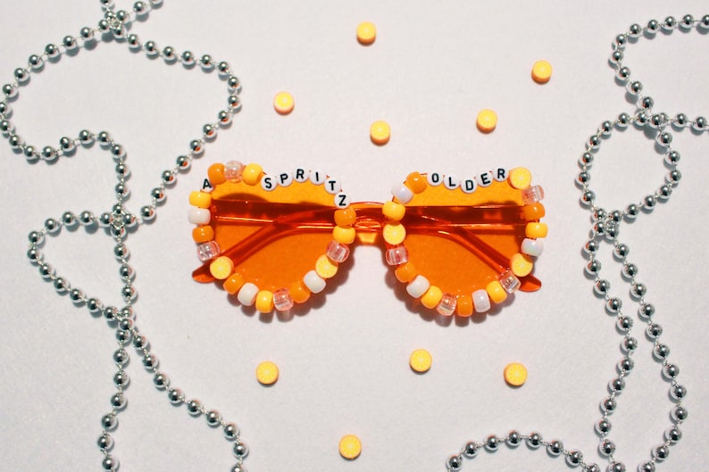 May include: Orange-tinted sunglasses with the text "A Spritz Older" spelled out in white letter beads. The frames are decorated with yellow, orange, and white beads. Silver beaded necklaces and yellow candies are scattered around the sunglasses.