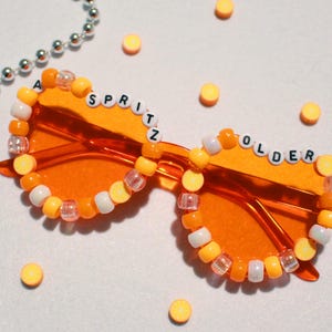 May include: Orange-tinted sunglasses with the words "A SPRITZ OLDER" spelled out in white letter beads. The frames are adorned with a beaded border of orange, white, and clear beads. Silver beaded necklaces and yellow circular objects are scattered around the sunglasses.