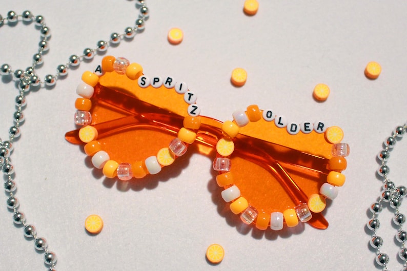 May include: Orange-tinted sunglasses with the words "A SPRITZ OLDER" spelled out in white letter beads. The frames are adorned with a beaded border of orange, white, and clear beads. Silver beaded necklaces and yellow circular objects are scattered around the sunglasses.
