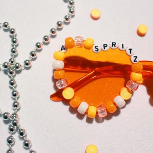 May include: Orange-tinted sunglasses with the word "SPRITZ" spelled out in white letter beads. The frames are decorated with orange, yellow, and white beads. A silver beaded necklace is in the background, along with small orange circle decorations.