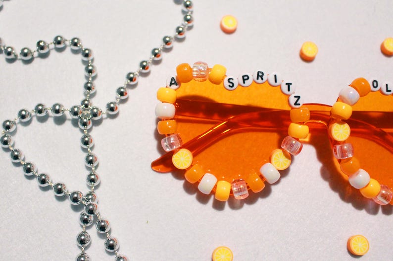 May include: Orange-tinted sunglasses with the word "SPRITZ" spelled out in white letter beads. The frames are decorated with orange, yellow, and white beads. A silver beaded necklace is in the background, along with small orange circle decorations.
