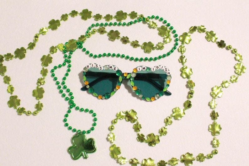 Peut inclure: Des lunettes de soleil vertes avec le texte "Kiss Me I'm Irish" sont entour&eacute;es de colliers de perles et de tr&egrave;fles verts. Les lunettes de soleil ont une monture en forme de c&oelig;ur et sont d&eacute;cor&eacute;es de gemmes vertes, oranges et transparentes.