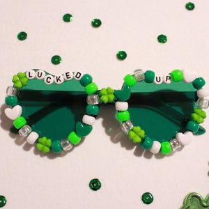 May include: Green sunglasses with the words "LUCKED UP" spelled out in white letters. The frames are decorated with green, white, and clear beads, including shamrock and heart shapes. Green sequins and a beaded necklace are also visible.