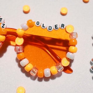 May include: Orange-tinted sunglasses with beaded embellishments spelling "SPRITZ" and "OLDER". The frames are adorned with yellow, white, and clear beads, some shaped like lemon slices. A silver beaded necklace is in the background.