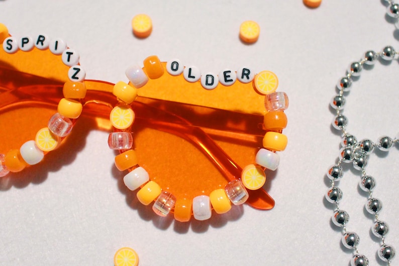 May include: Orange-tinted sunglasses with beaded embellishments spelling "SPRITZ" and "OLDER". The frames are adorned with yellow, white, and clear beads, some shaped like lemon slices. A silver beaded necklace is in the background.