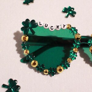 K&ouml;nnte beinhalten: Gr&uuml;ne Sonnenbrille, verziert mit Kleebl&auml;ttern und goldfarbenen Verzierungen. Das Wort "LUCKY" ist mit wei&szlig;en Buchstabenperlen auf der Oberseite des linken Glases geschrieben. Verstreute Kleeblatt-Konfetti umgeben die Brille.