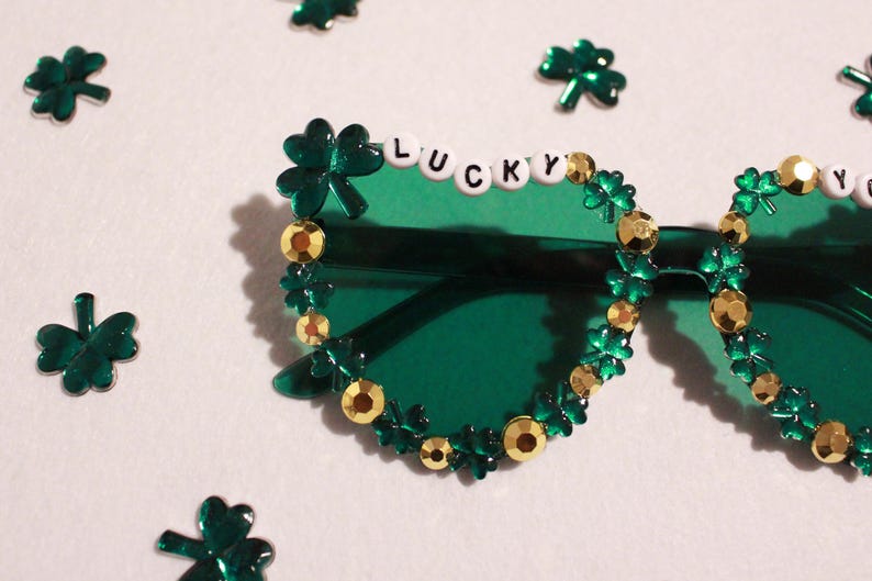 K&ouml;nnte beinhalten: Gr&uuml;ne Sonnenbrille, verziert mit Kleebl&auml;ttern und goldfarbenen Verzierungen. Das Wort "LUCKY" ist mit wei&szlig;en Buchstabenperlen auf der Oberseite des linken Glases geschrieben. Verstreute Kleeblatt-Konfetti umgeben die Brille.