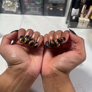 Puede incluir: Uñas ovaladas con un diseño brillante de ojo de gato en tonos marrón y dorado. Las uñas son largas y están bien cuidadas, con una base oscura y un efecto brillante. Las manos se muestran sobre un fondo neutro.