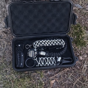 Pode incluir: Uma caixa preta aberta contendo uma faca dobrável preta, uma pulseira de paracord preta e branca, um chaveiro e um mosquetão. Os itens estão dispostos sobre uma base de espuma preta.
