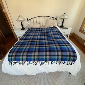 Puede incluir: Una manta a cuadros azules, negros y verdes con flecos cubre una cama con sábanas blancas. La manta es el punto focal, con un patrón repetitivo. La cama está en una habitación con mesitas de noche y lámparas blancas.