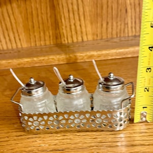 Pode incluir: Um conjunto de condimentos vintage com três pequenos frascos de vidro, tampas e colheres prateadas, numa bandeja decorativa de metal prateado. Os frascos têm um padrão texturizado. O conjunto tem aproximadamente 7,6 cm de comprimento.