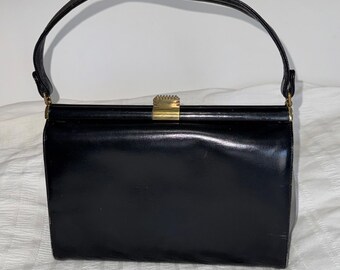 Puede incluir: Un bolso negro rectangular con un asa curva. El bolso tiene un acabado brillante, similar al charol, y un cierre dorado. El asa está sujeta a la parte superior del bolso con herrajes dorados. El bolso está sobre una superficie blanca.