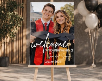 May include: A graduation party welcome sign on a wooden easel. The sign features a photo of a smiling couple in graduation attire, with the word "welcome" in cursive script. Text below reads "To the graduation party of Tom and Sandy, Class of 2026."