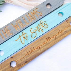 May include: Three wooden signs in gray, teal, and brown, each with a circular hole and engraved text. The gray sign reads "THE WILLIAMS", the teal sign reads "the Smiths", and the brown sign reads "Ashley & Miguel".